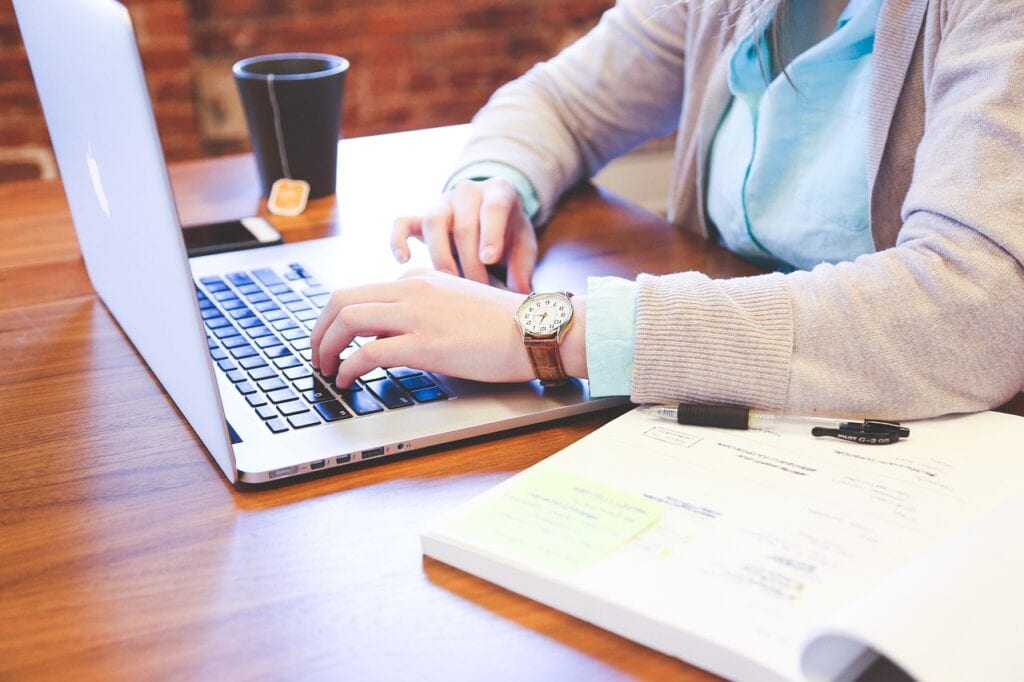 Pessoa com relógio digitando em um notebook sobre uma mesa com um livro ao lado e uma xícara. 