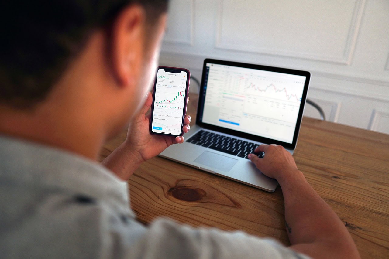 Homem analisando gráficos de investimentos em celular e laptop, representando ações e finanças pessoais.