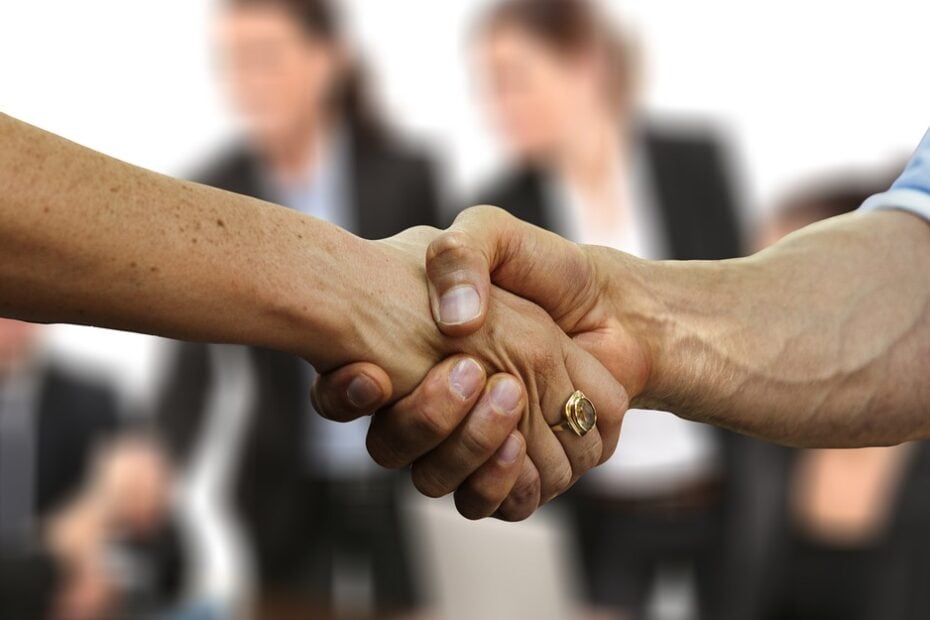 A imagem mostra duas mãos se cumprimentando, simbolizando um acordo ou parceria, com pessoas ao fundo representando um ambiente de negócios.