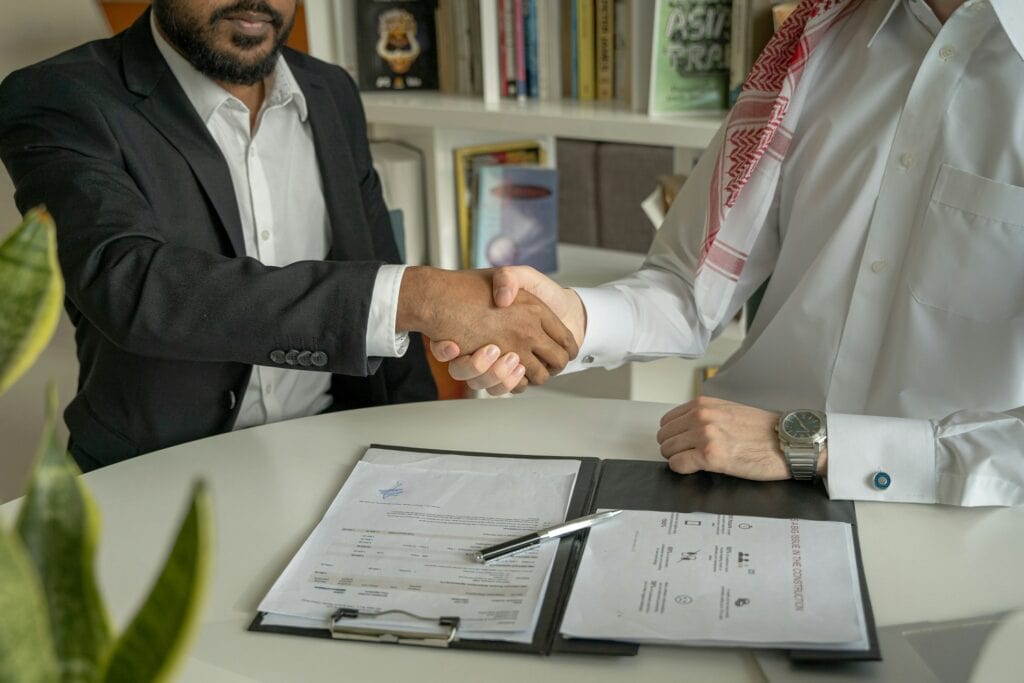Homem de terno e homem com camisa branca apertando as mãos em acordo de negócios em escritório, simbolizando parceria e sucesso empresarial.