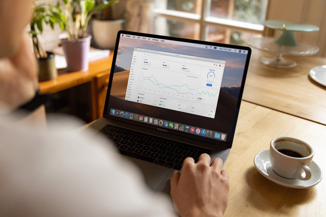 Pessoa usando laptop com gráficos financeiros na tela, acompanhando dados de desempenho. Xícara de café e ambiente aconchegante.