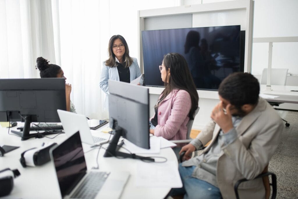 Reunião em escritório moderno com quatro pessoas conversando sobre projetos e ideias. A líder discute com a equipe frente a computadores.